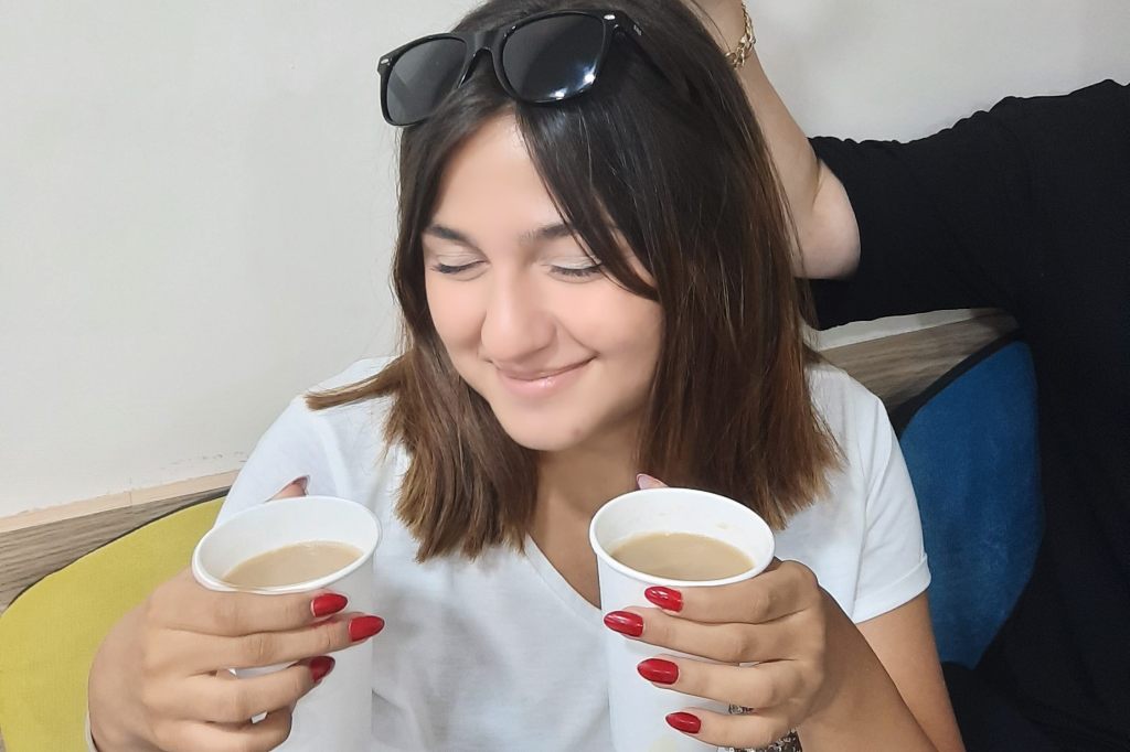Elif holding two cups of coffee in class, smiling during roll call where her coffee became a fun ritual with her teacher.